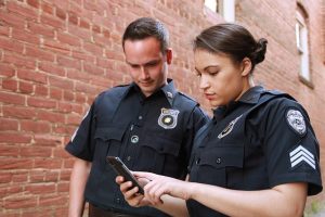 Image is a male and female police officer standing in front of a brick wall looking at a phone.