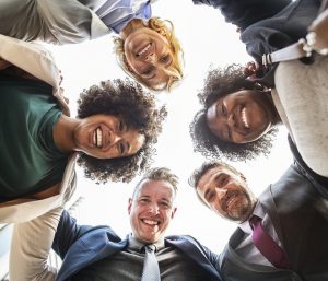 Image is a group of men and women in a circle smiling, looking down at a camera on the floor in the middle of them.