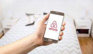 Image is of a person holding a phone with the airbnb app open, while standing in a bedroom.