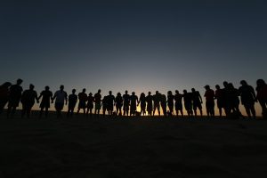 Image is a line of people holding hands, backlit and silhouetted by the setting sun.